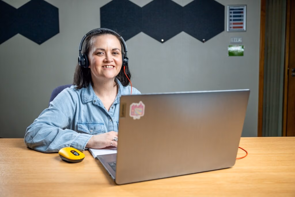 Claire Cross in front of a laptop and wearing a headset, looking towards the camera.