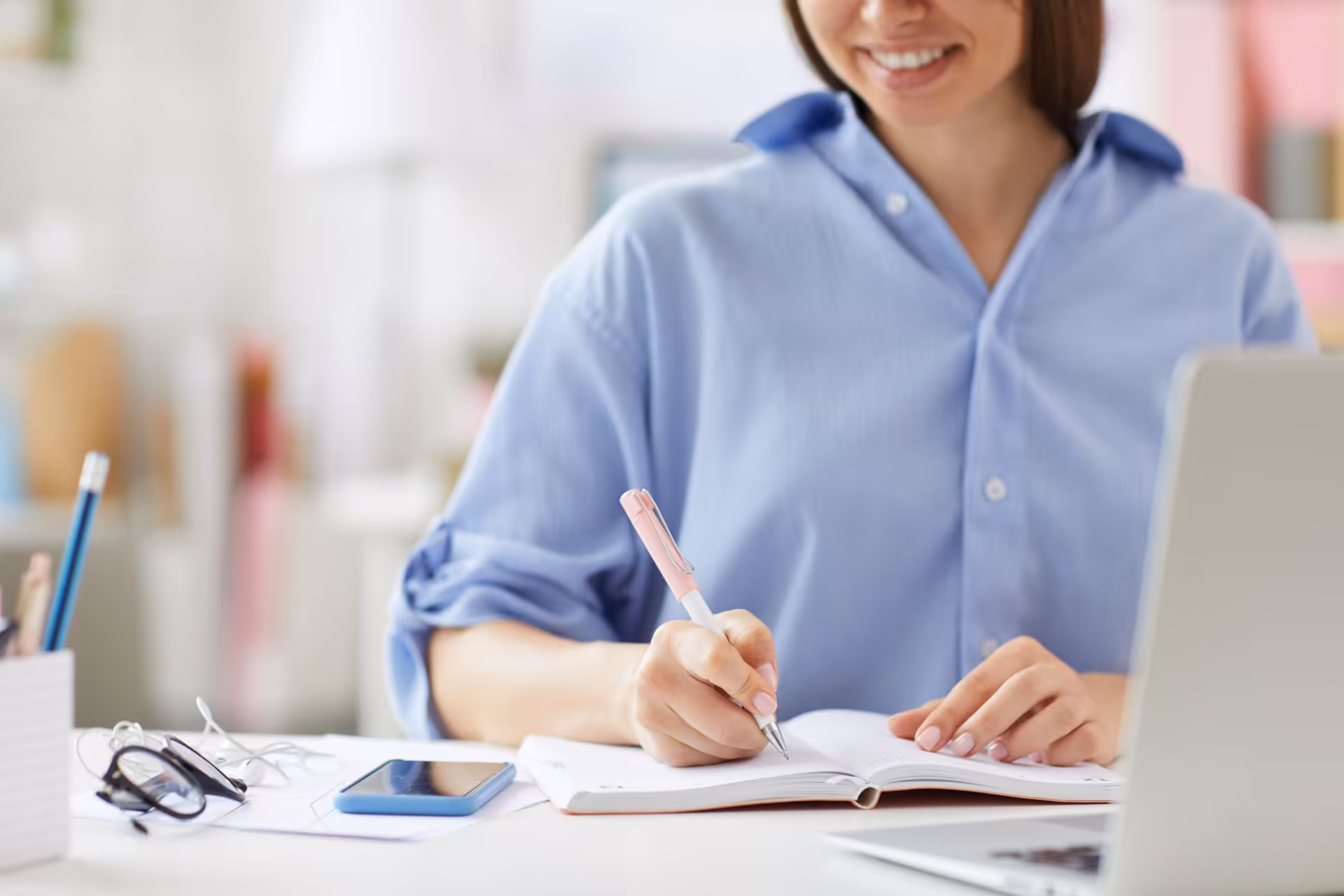 A young woman in a blue shirt sitting at a desk. She is writing a plan for the day in a book.